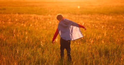 A Child in the Costume of a Superhero in a Red Cloak Runs Across the Green Lawn Against the Backdrop