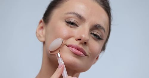 Close Up Portrait of Young Woman Using Jade Facial Roller for Skin Care Beauty Treatment on Gray