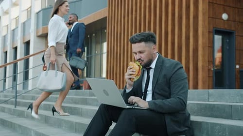 Slow Motion of Male Office Worker Using Laptop and Eating Sandwich Sitting on Stairs Outside Office