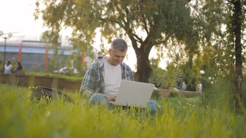 Mann arbeitet hinter einem Laptop und sitzt am sonnigen Tag im Gras