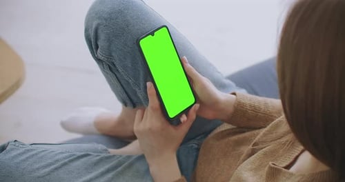 Woman at Home Lying on a Couch Using Smartphone with Green Mock-up Screen, Doing Swiping, Scrolling