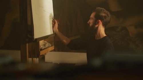 Bearded Man Drawing on Easel in Dimly Lit Studio