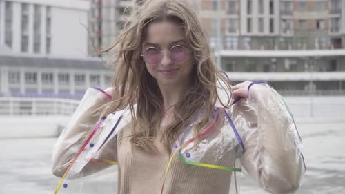 Porträt eines positiven süßen Mädchens mit Sonnenbrille, das einen transparenten Regenmantel anzieht und lacht. Charming