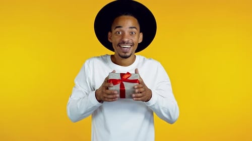Young Afro Man Holding Gift Box with Bow, He Wonders What Is Inside. Yellow Wall Background. African