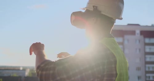 Engineer Builder on the Roof of the Building at Sunset Stands in VR Glasses and Moves His Hands
