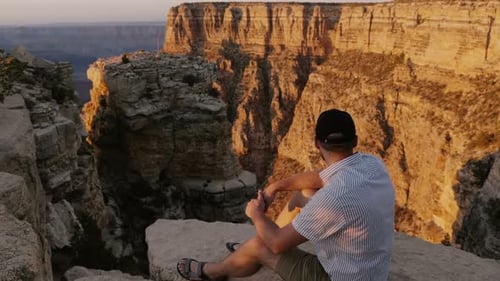 Hochwinkelkamera bewegt sich über glücklichen jungen erwachsenen Touristen, der auf einem großen Felsen sitzt und den Sonnenuntergang beobachtet