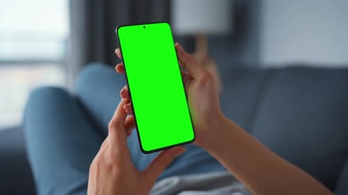 Woman at Home Lying on a Sofa and Using Smartphone with Green Mock-up Screen in Vertical Mode. Girl