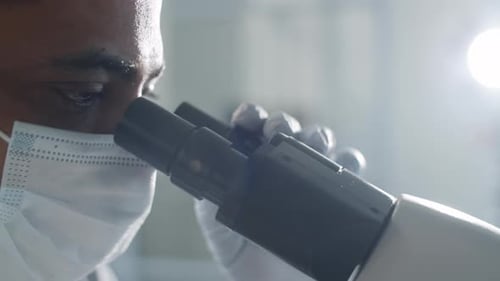Scientist in Mask and Gloves Looking through Microscope