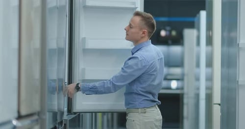 A Young Man in a Blue Shirt To Open the Door of the Refrigerator in the Appliances Store and Compare