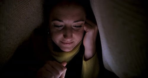 Woman lying on the bed under the blanket and using smartphone navigating on s