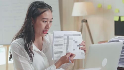 Close Up Of Asian Woman With Documents Explaining Work On Paper While Having Video Call On Laptop