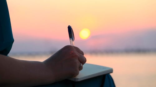 Woman Writes in Notebook by Water at Sunset