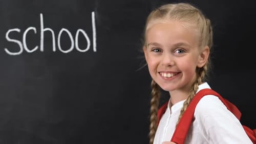 Little Girl With Rucksack Smiling at Camera, School Word Written on Blackboard