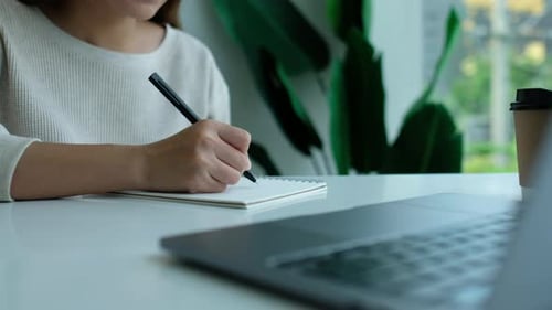 Woman Writing in Notebook at Desk with Laptop