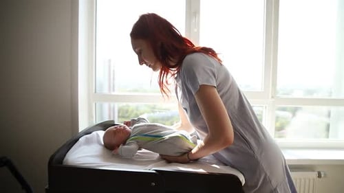 Ginger Woman Mother Puts Her Baby on the Baby Cot and Patting His Stomach