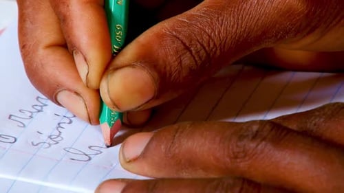 Extreme close up of an African person's hands as they write on lined white paper in a classroom in A