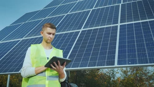 Ein junger Ingenieur für Arbeitskleidung arbeitet mit einem Tablet in der Nähe eines großen Solarpanels.
