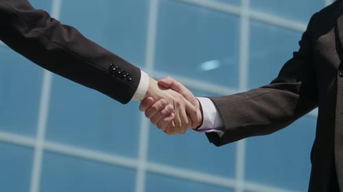 Male strong handshake of people in classic black suits, against the background of a modern office