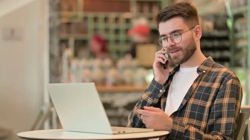 Young Man with Laptop Talking on Smartphone in Cafe