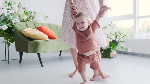 Happy little baby learns to walk with the help of mother at home. Bedroom interior with green sofa a