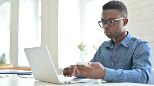 Focused Young African Man Using Laptop and Smartphone in Office