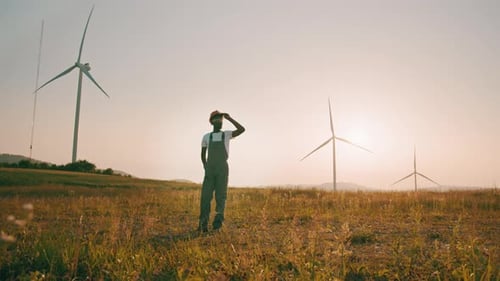 Hombre afroamericano de pie en el campo con enormes molinos de viento durante el atardecer de verano