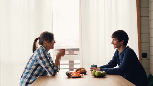 Side View of Man and Woman Drinking Tea and Talking Sitting at Table in Kitchen Together Enjoying