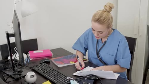 Young Beautiful Blond Female Doctor in Blue Coat, Working at Desk Using Computer and Doing Paperwork