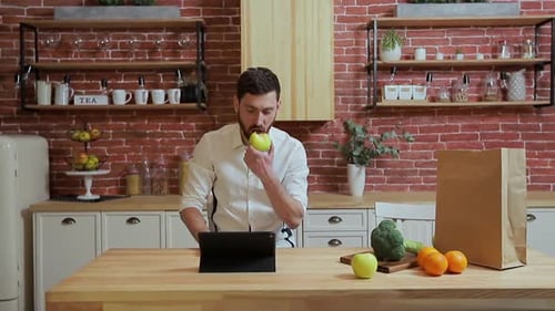 Man Using Tablet Eats Apple in Kitchen
