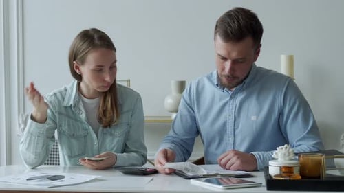 Young Married Couple Sitting at Living Room Table Studying Papers Makes Calculations on a Calculator