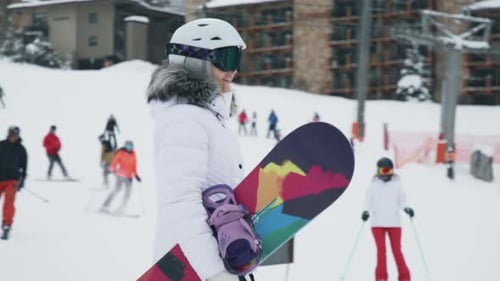 Frau mit Snowboard geht am verschneiten Berg entlang und Skifahrer fahren den Hang hinunter