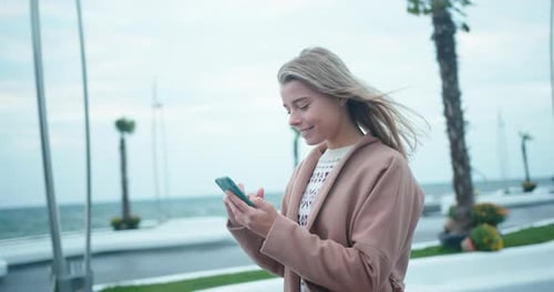 Young Woman Strolls Along Embankment and Chats Using Phone
