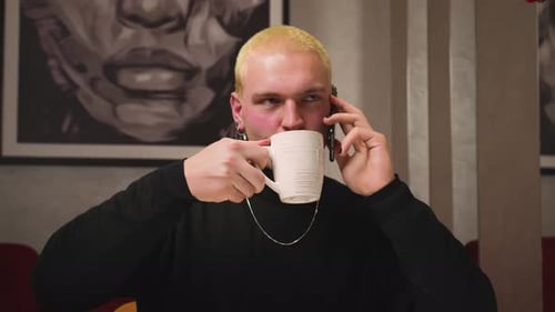 Hübscher lächelnder homosexueller Mann mit gelben Haaren und Maniküre telefoniert und Kaffee trinkt