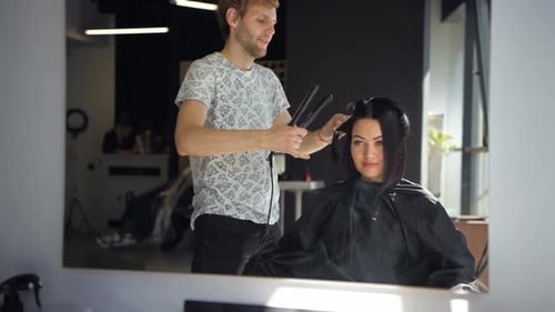 El peluquero alisa el cabello castaño para una mujer usando una plancha de pelo en el salón de belleza