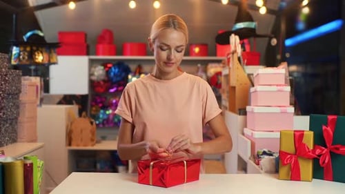 Front View of Young Woman Worker Holding and Using Golden Ribbon to Tie Bow for Red Wrapped Gift Box
