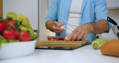 Hombre que da sal y pimienta a los filetes en la tabla de cortar antes de asarlos en la cocina moderna