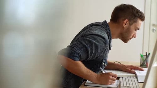Side View of University Male Student in Casual Clothes Studying Taking Notes From Book While Sitting