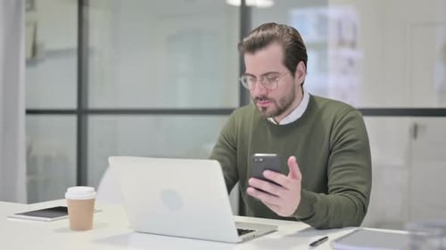 Young Businessman Using Smartphone While Using Laptop in Office