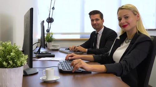 Two office workers, man and woman, work on computers and smile at the camera
