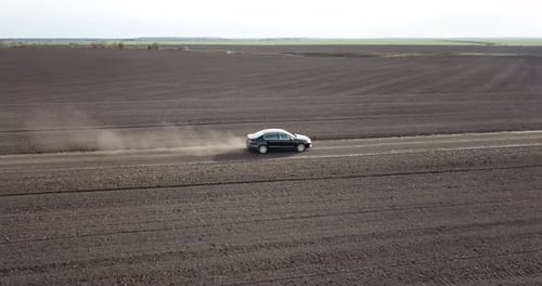 The Car is Racing Along the Road in the Countryside and Blowing a Dusty View From