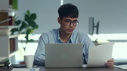 Young professional business male using computer looking information paper at office.