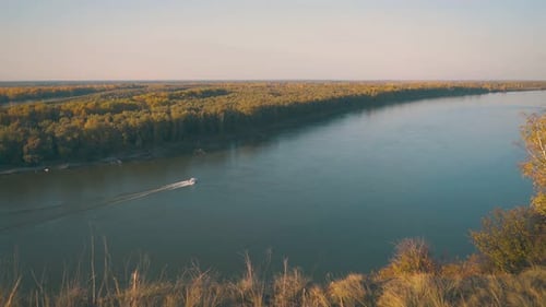 Motorboat Sails Along Calm River at Dense Forest in Autumn