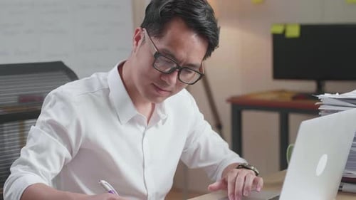 Close Up Of Asian Man Looking At Laptop And Taking Note While Working With Documents At The Office