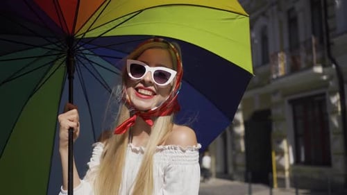 Cute Young Woman Looking at the Camera Holding Multicolored Umbrella Standing on the Street