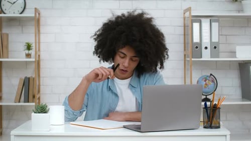Nachdenklicher Student, der drinnen am Laptop sitzt und lernt, Notizen zu machen