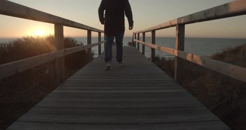 Person Walking on Boardwalk Towards Ocean at Sunset