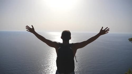 Unrecognizable Hiker with Backpack Standing on Edge of Mountain and Victoriously Raising Hands