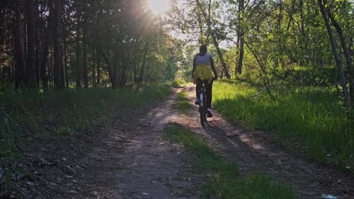 Young Woman on a Bicycle Rides Along Green Forest Path in Sunny Summer Day