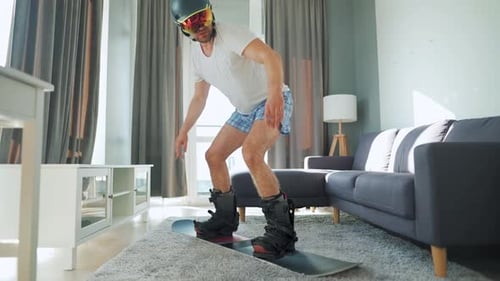 Vídeo divertido. Homem de bermuda e camiseta retrata snowboard em um tapete em um quarto aconchegante. Esperando por