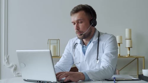Porträt eines Arztes im weißen Kittel, der bei Videoanruf im Headset auf Laptop spricht, der im Schrank sitzt